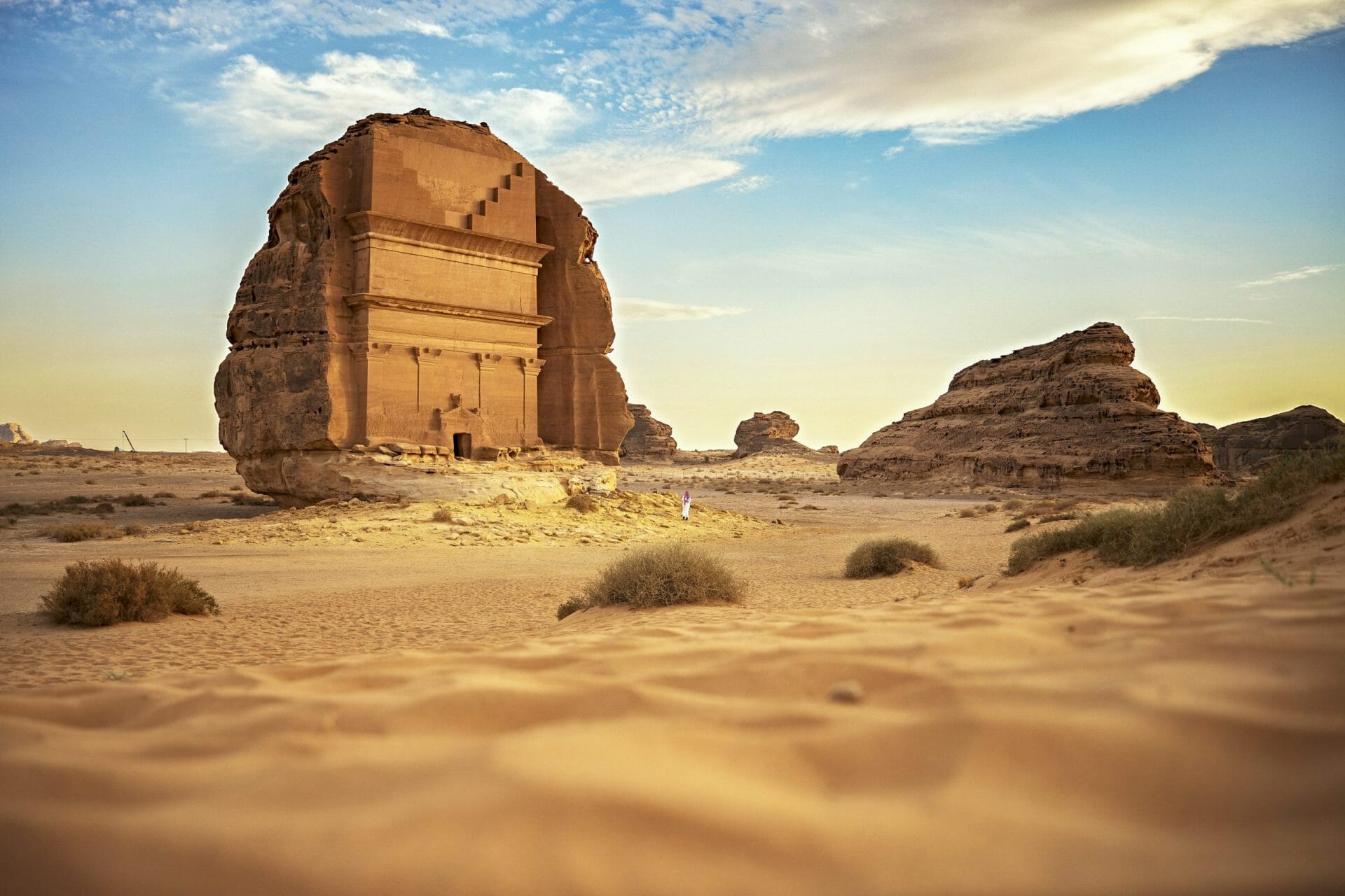 Surface level view from sand dune with focus on background Tomb of Lihyan, son of Kuza, iconic burial chamber cut into existing rock formation.