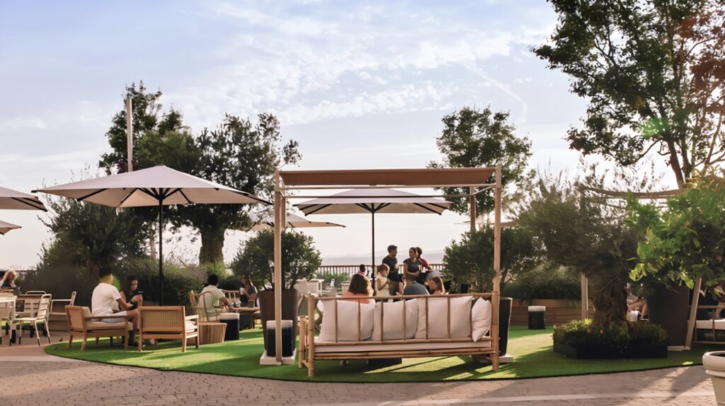 Group of people casually chatting around a table in a sunny outdoor lounge with beige sofas and large umbrellas under a pergola-like frame.