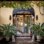 Entrance to a stylish building labeled The Apartment, with a black canopy, potted palm plants, and a small staircase leading inside.
