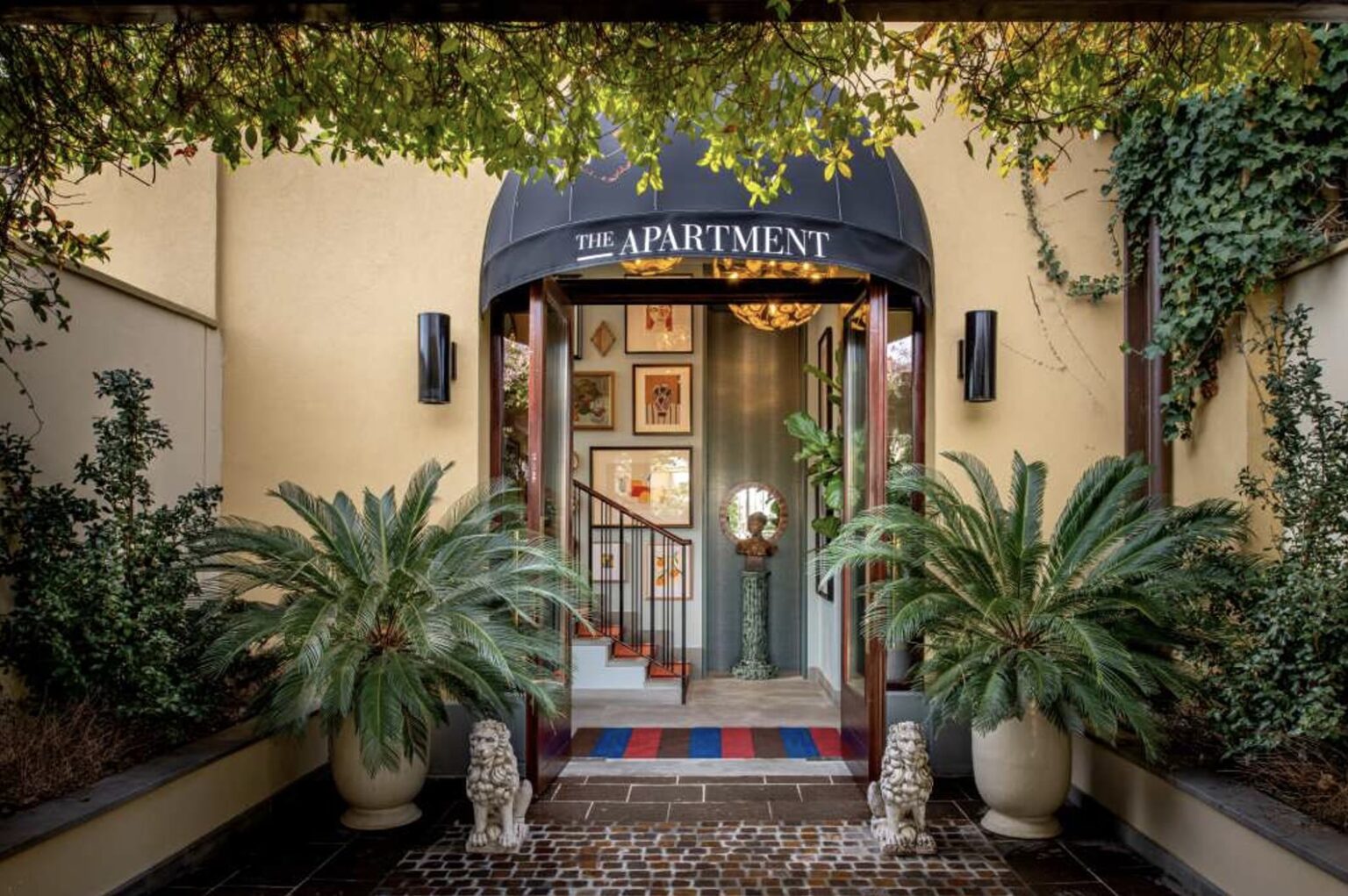 Entrance to a stylish building labeled The Apartment, with a black canopy, potted palm plants, and a small staircase leading inside.