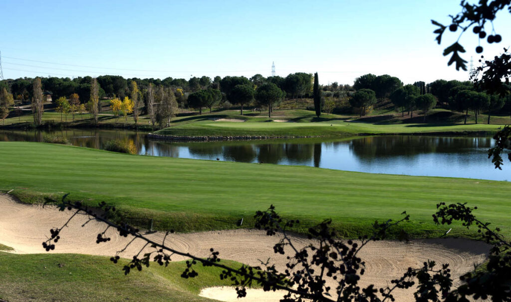Well-manicured golf course with a green fairway, a sandy bunker in the foreground, and a calm lake reflecting trees under a clear blue sky.