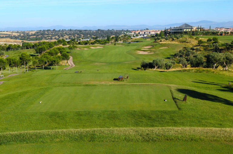 Well-manicured golf tee in the foreground with a wide fairway, sand bunkers, and trees stretching toward a distant clubhouse on a sunny day.