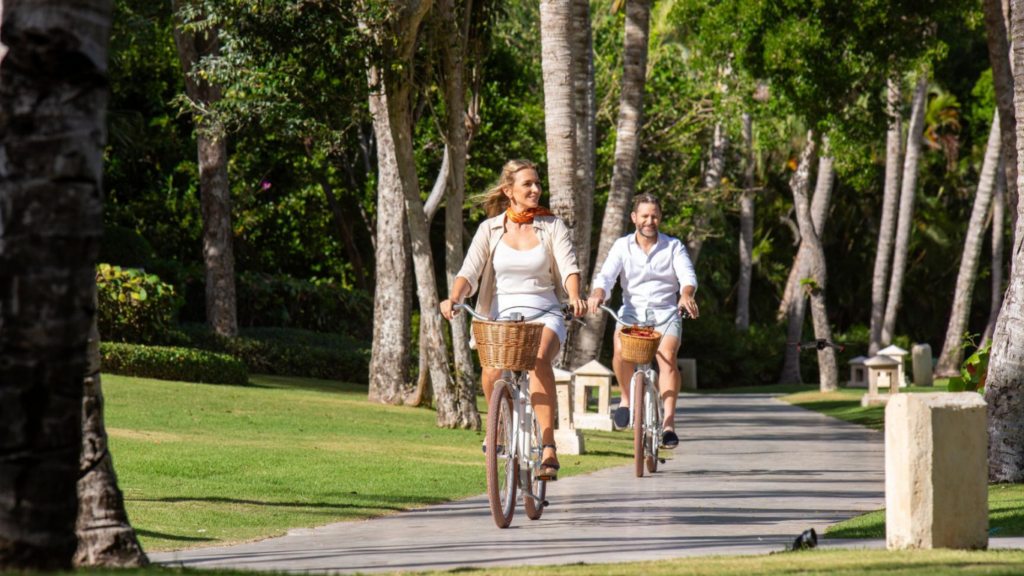 A couple rides vintage bikes with baskets along a tree-lined park path left to right in bright sunshine.