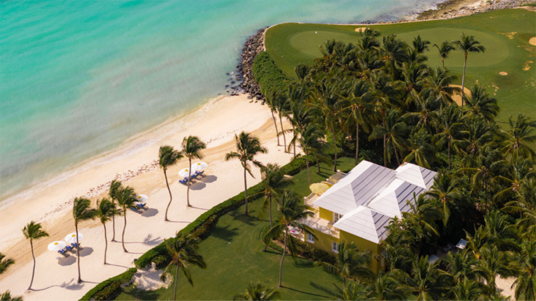 Aerial view of a tropical beach resort: turquoise water, white sand with lounge chairs and umbrellas, and a yellow building surrounded by palm trees.