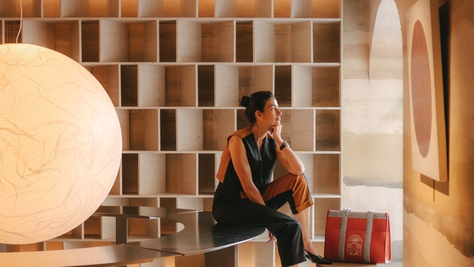 Woman sitting thoughtfully in a modern showroom, with a large white spherical lamp on the left and a red gift bag on the floor nearby.