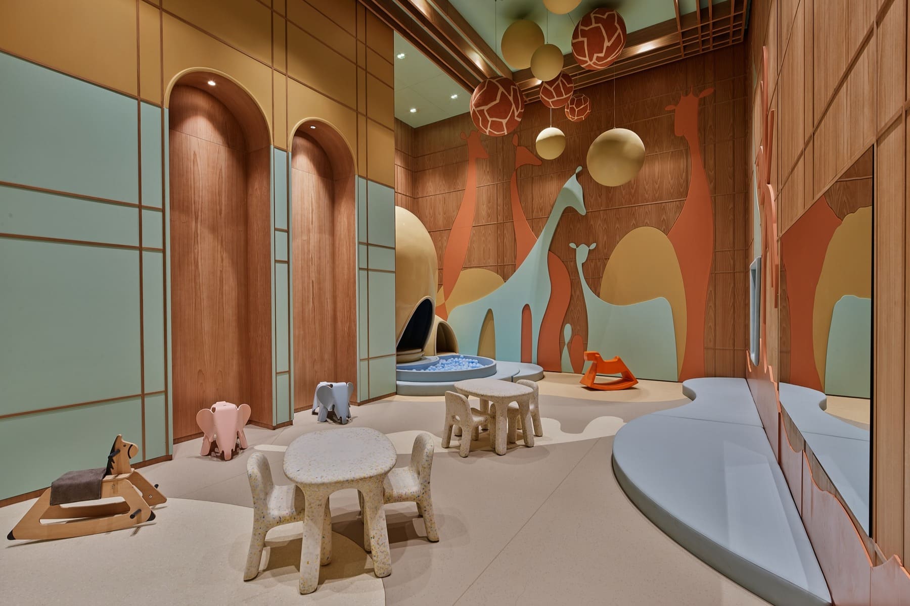 Colorful children's playroom with pastel orange and blue walls, wall mural of giraffes, and soft foam seating around a shallow ball pit and toy animals.