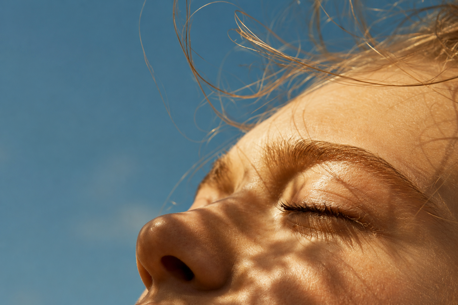 Close-up of a person’s sunlit face with eyes closed against a clear blue sky.