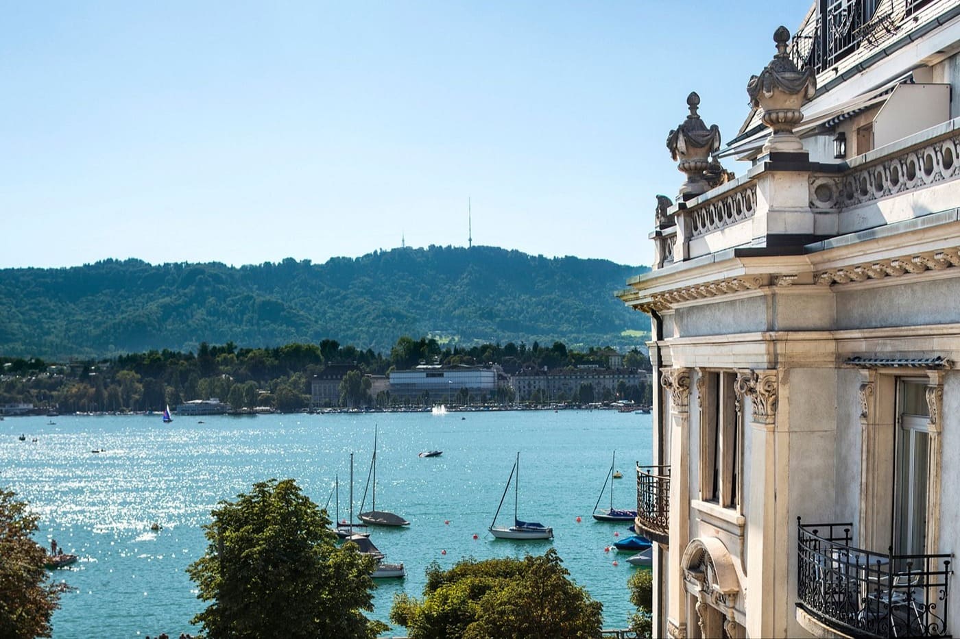 View across a blue lake with sailboats and a tree-lined shore, framed by an ornate stone building on the right.