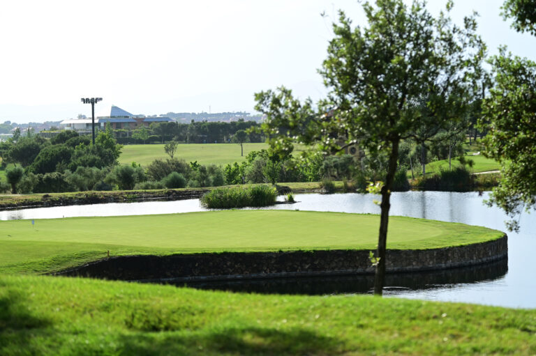 Lush golf course with a green fairway, a water hazard along a stone edge, and trees in the foreground; buildings in the distance under a clear sky.