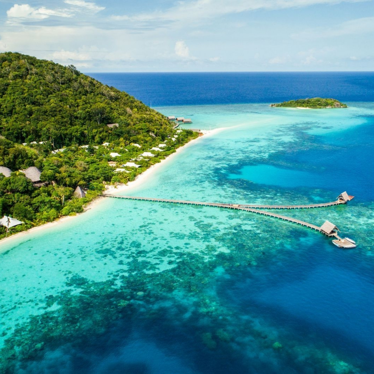 Tropical island resort with a curved wooden pier extending over turquoise water to overwater huts beside a forested shore.