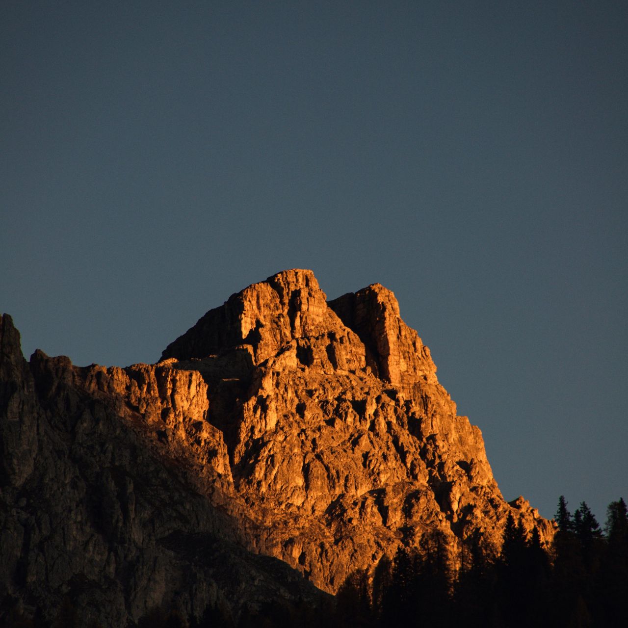 Rugged mountain peak bathed in warm sunset light with dark blue sky above and pine trees at the base