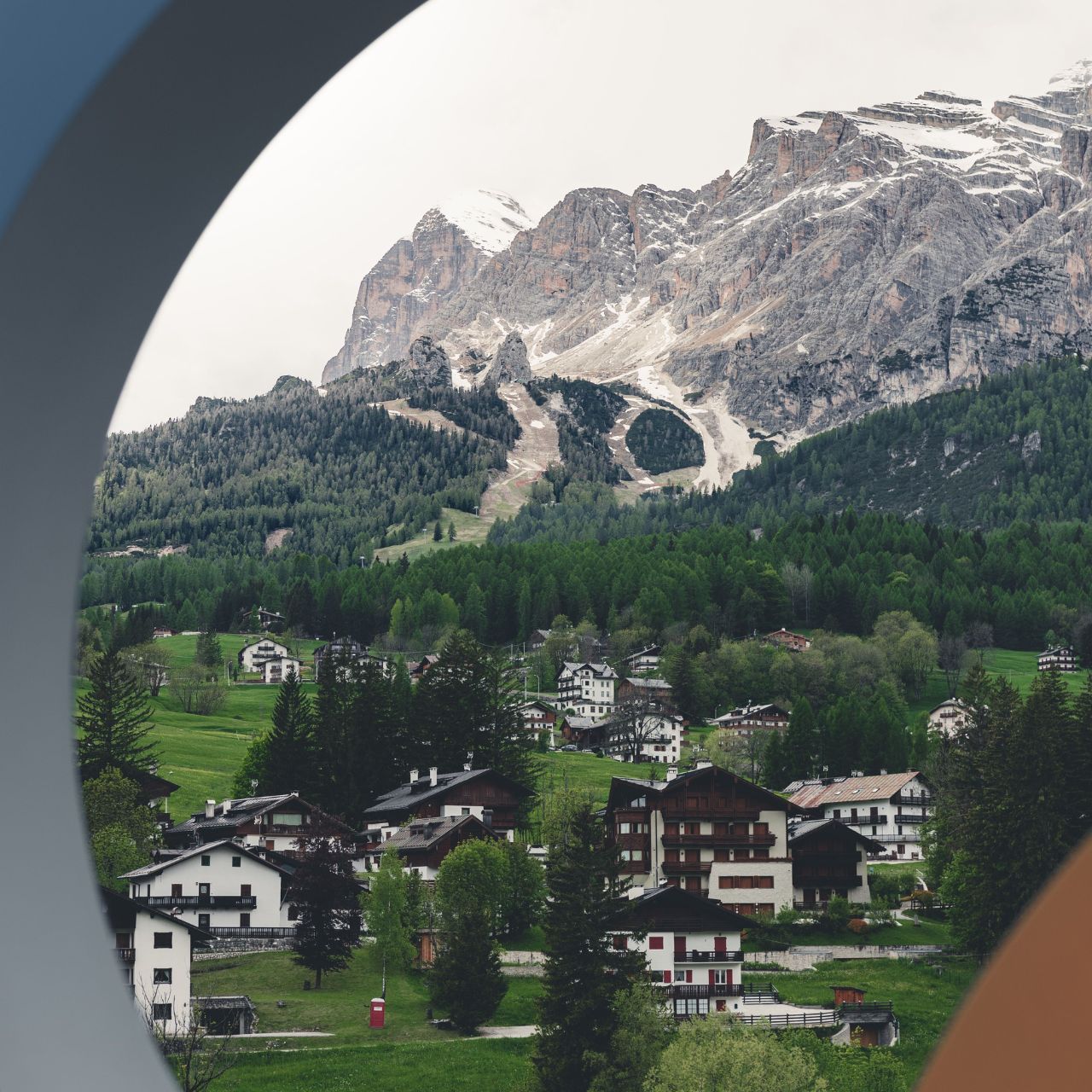 Alpine village with white houses and green fields at the base of snow-dusted mountains, viewed through a circular frame.