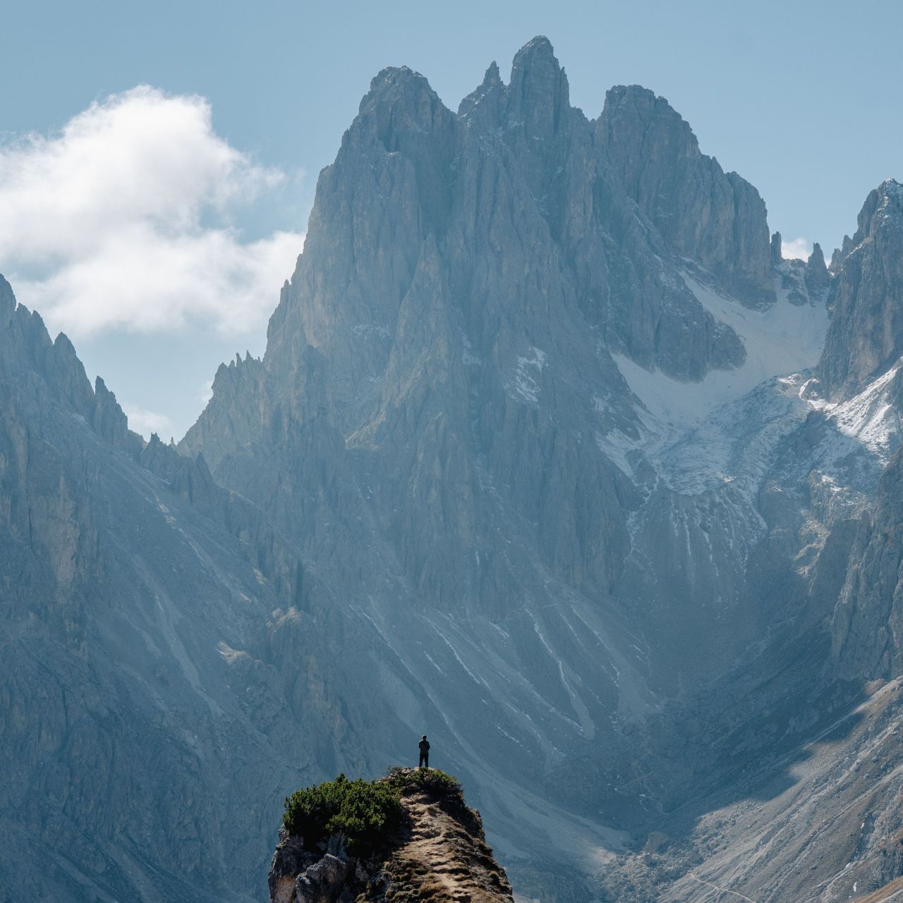 Person standing on a rocky ledge overlooking towering snow-dusted mountains under a blue sky.