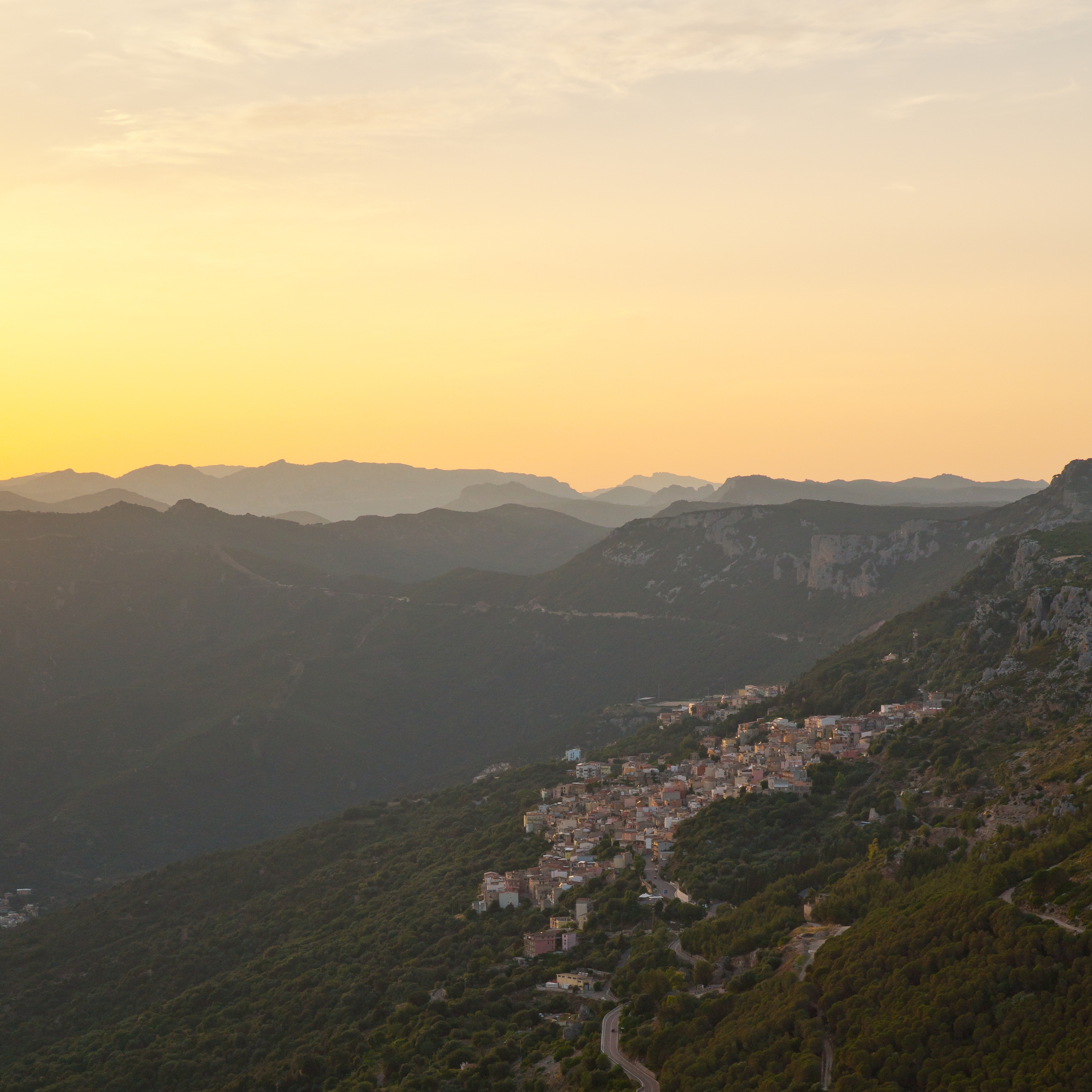 Sunset over a mountainous landscape with a hillside town and a winding road along the slope at dusk.