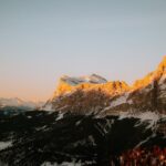 Snow-covered alpine peaks bathed in warm sunset light with a dark, forested valley below