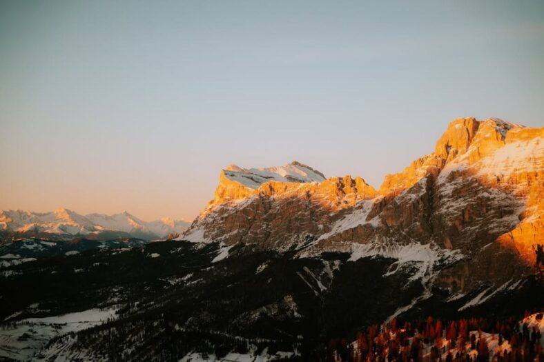Snow-covered alpine peaks bathed in warm sunset light with a dark, forested valley below