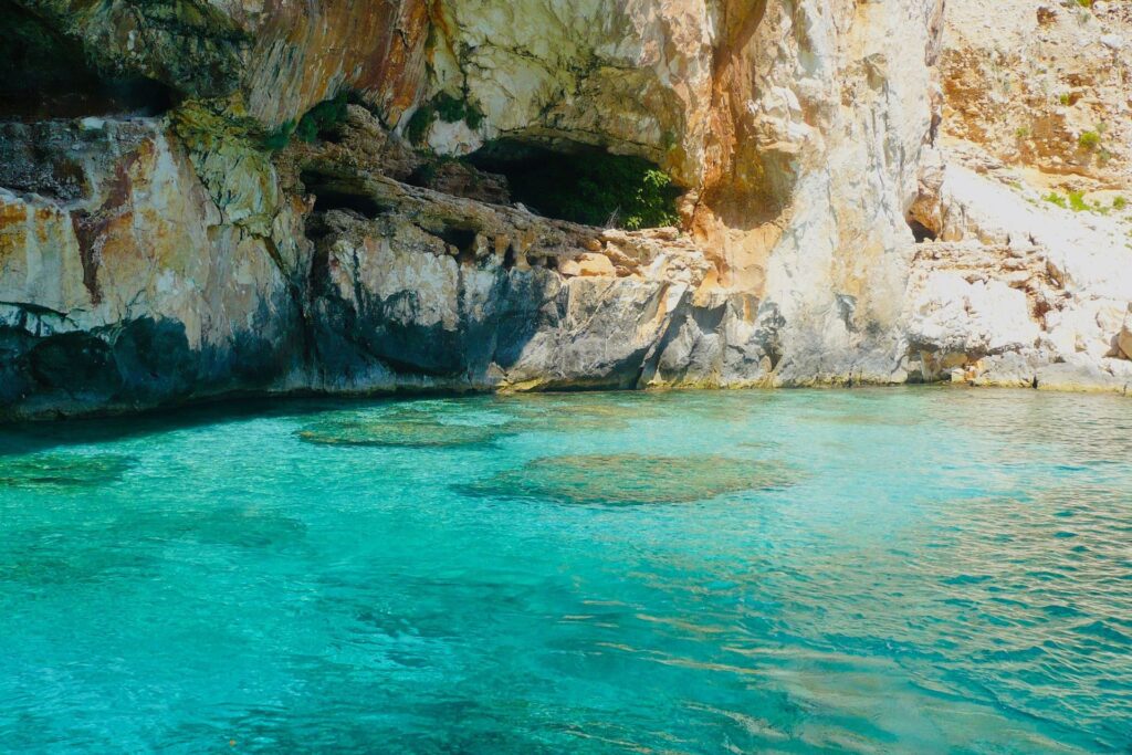 Turquoise coastal water lapping against rugged light-brown cliffs and a rocky cave opening in the rock face