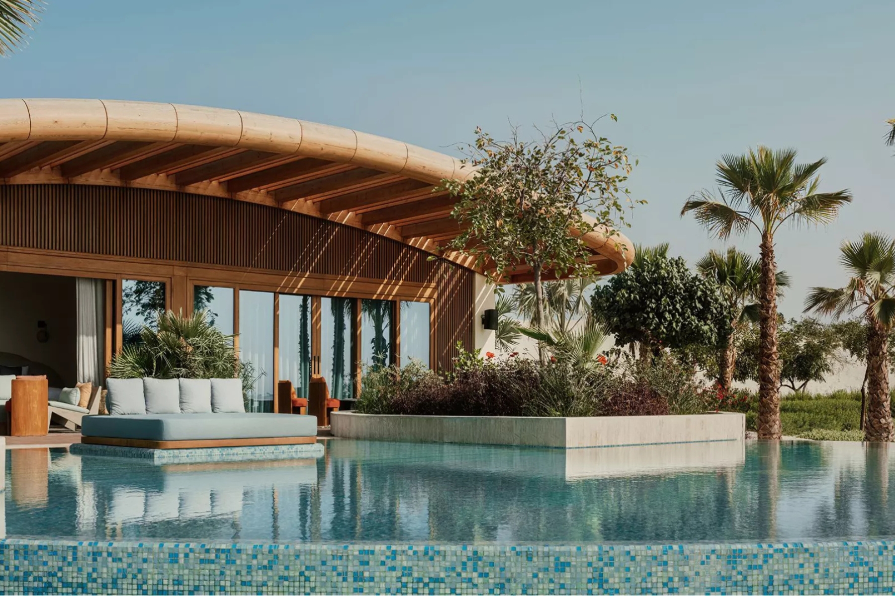 Luxurious poolside lounge under a curved wooden canopy with glass doors and palm trees nearby, sunny outdoor setting.