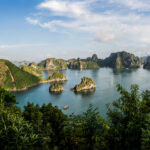 Calm blue bay with numerous green, rocky islands rising from the water under a bright sky; foreground frames with lush green foliage.