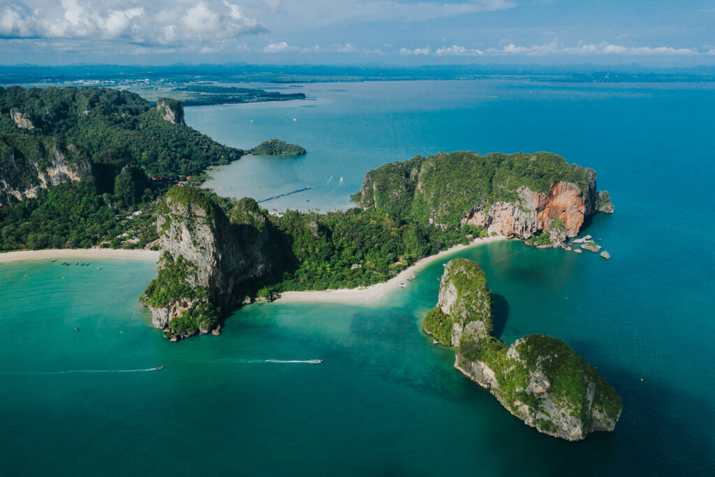 Aerial view of a tropical coastline with limestone cliffs, dense green forest, white sandy beaches, and turquoise waters.