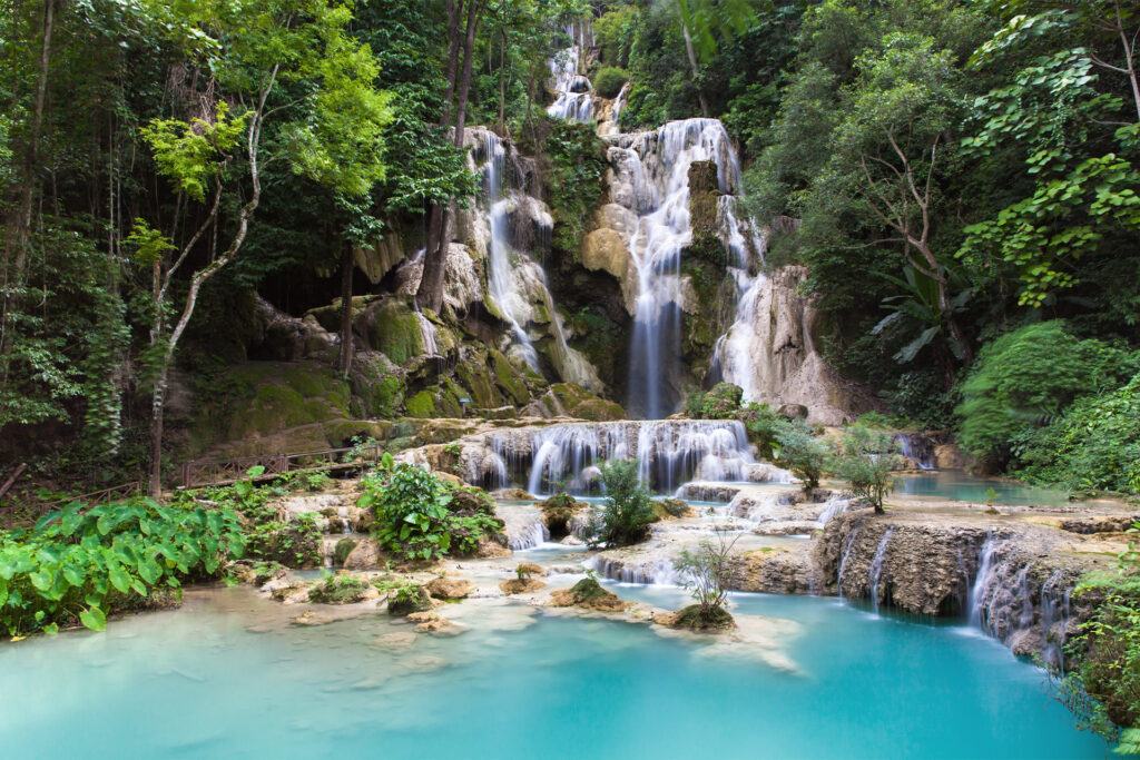 Tiered waterfall cascading over mossy rocks into turquoise pools in a dense green jungle, surrounded by tropical trees.