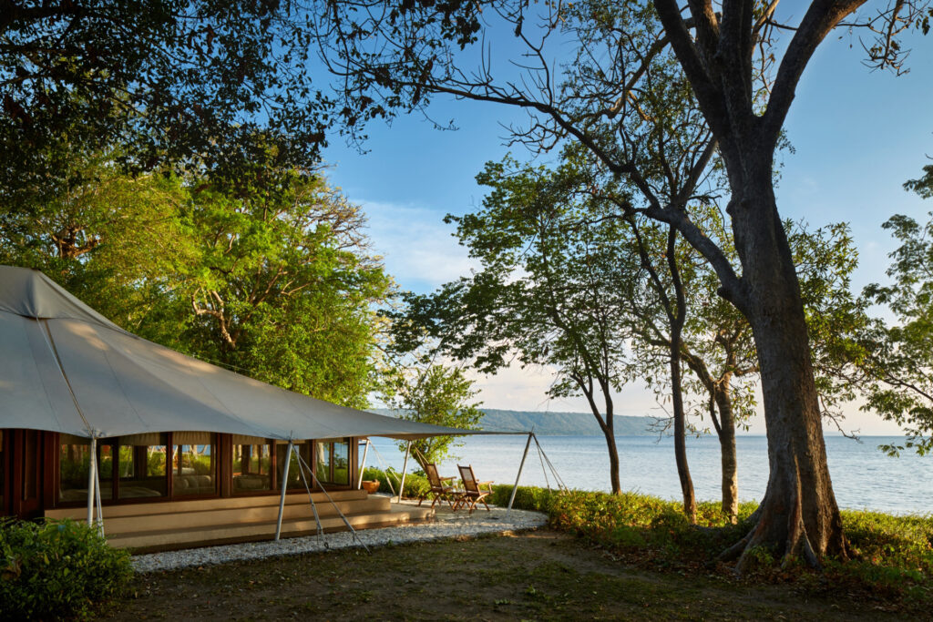 White canvas tent at a shady lakeside campsite with trees and chairs facing the water