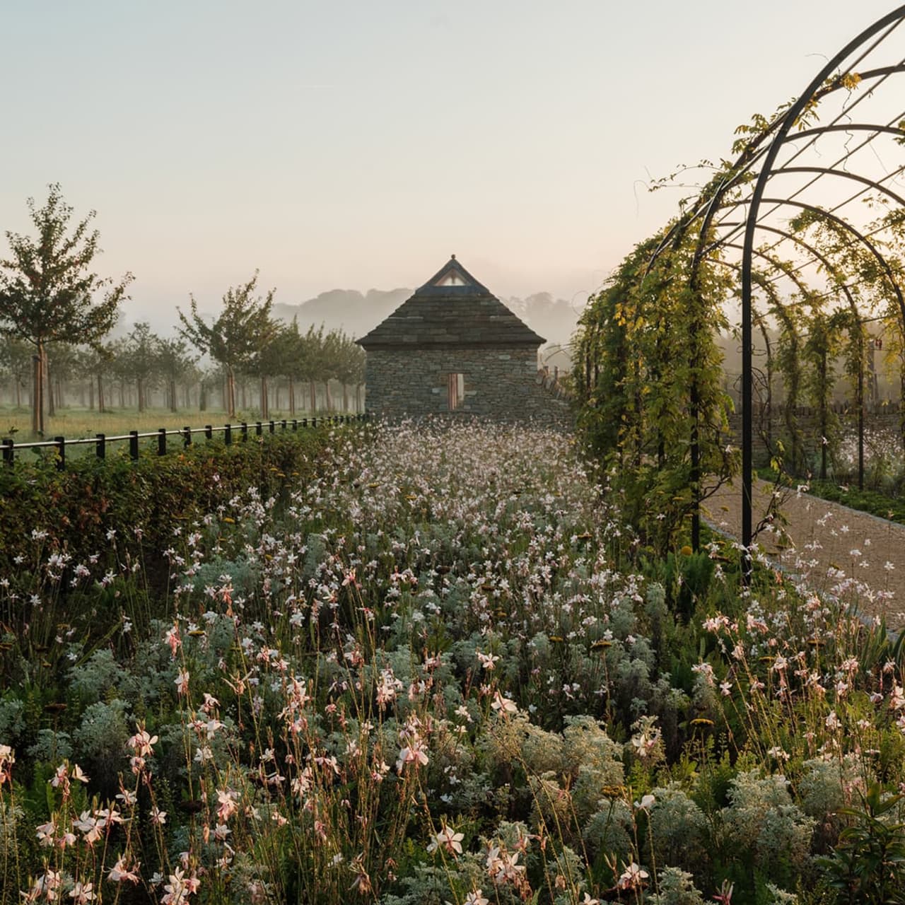 Stone cottage at the center of a flowering garden, with a vine-covered arched trellis on the right and soft morning mist in the background.