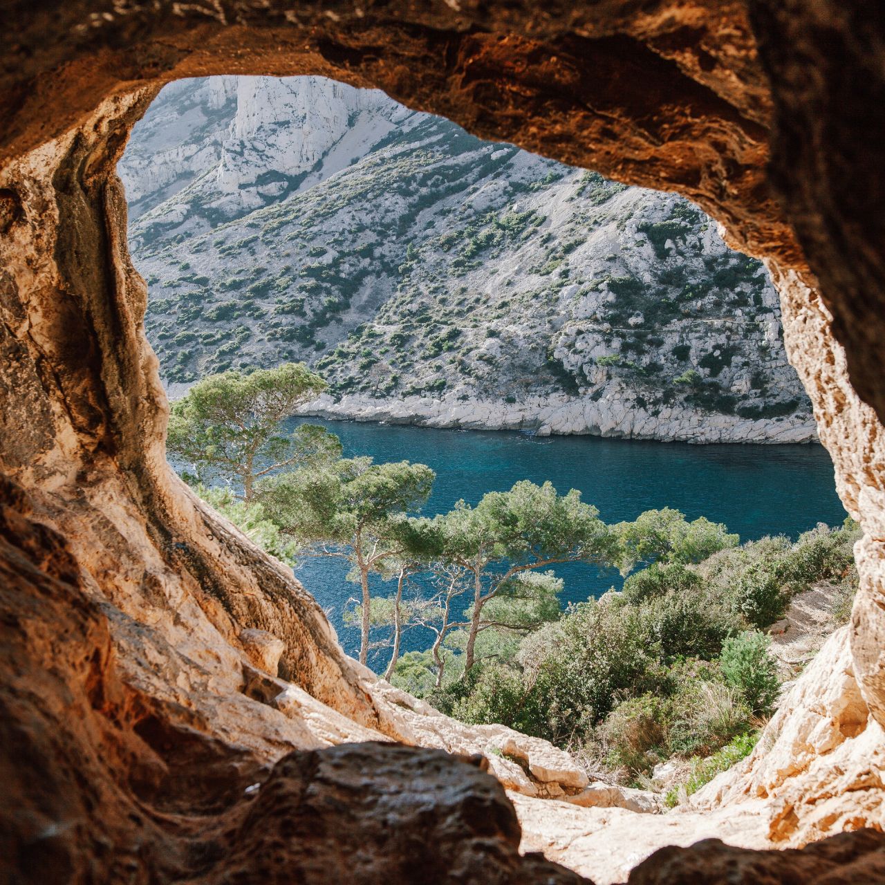View through a rocky cave opening onto a turquoise coastal inlet with steep cliffs and trees along the shore.