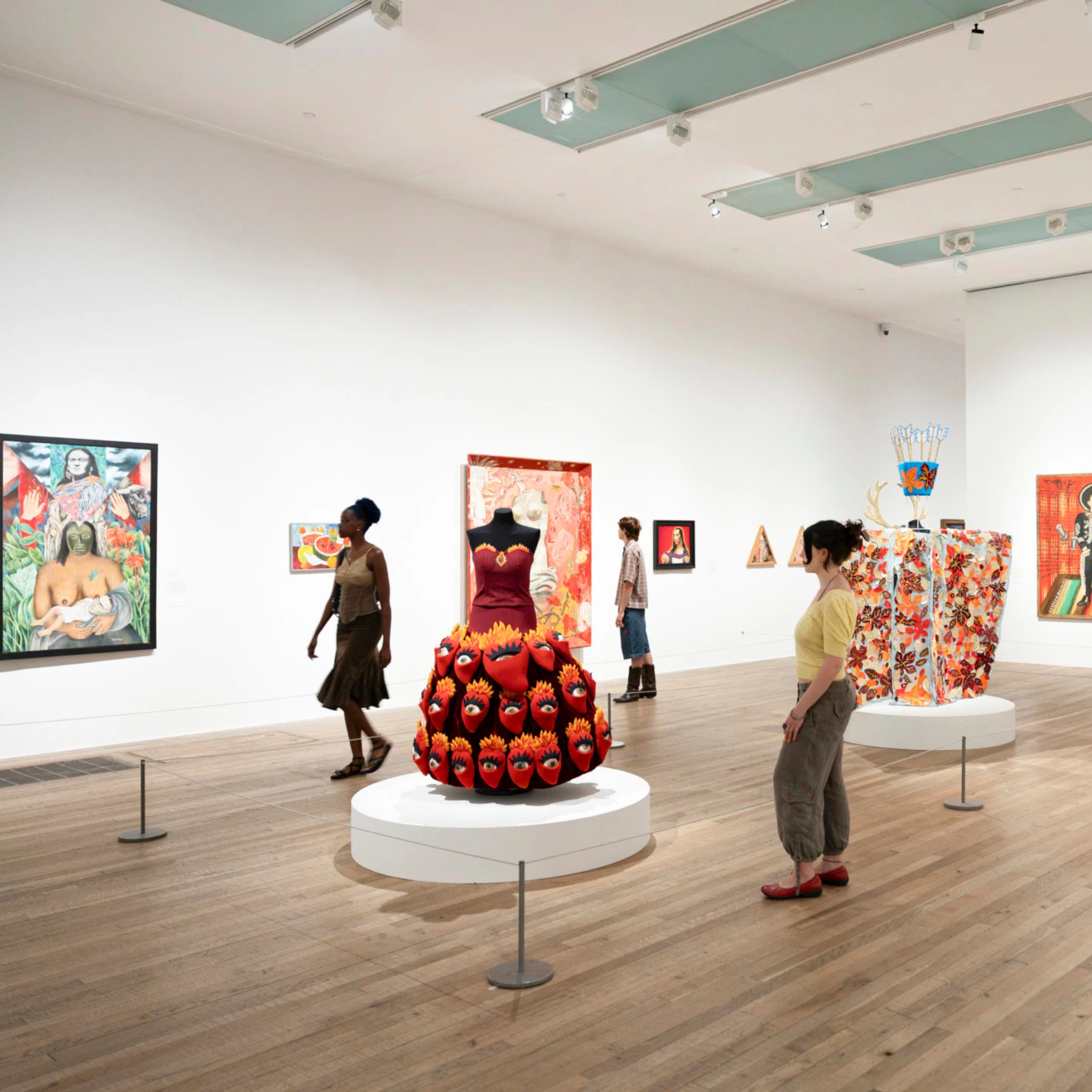 Red, multi-faceted sculpture on a pedestal in a bright gallery, with visitors viewing wall-mounted artworks around it.