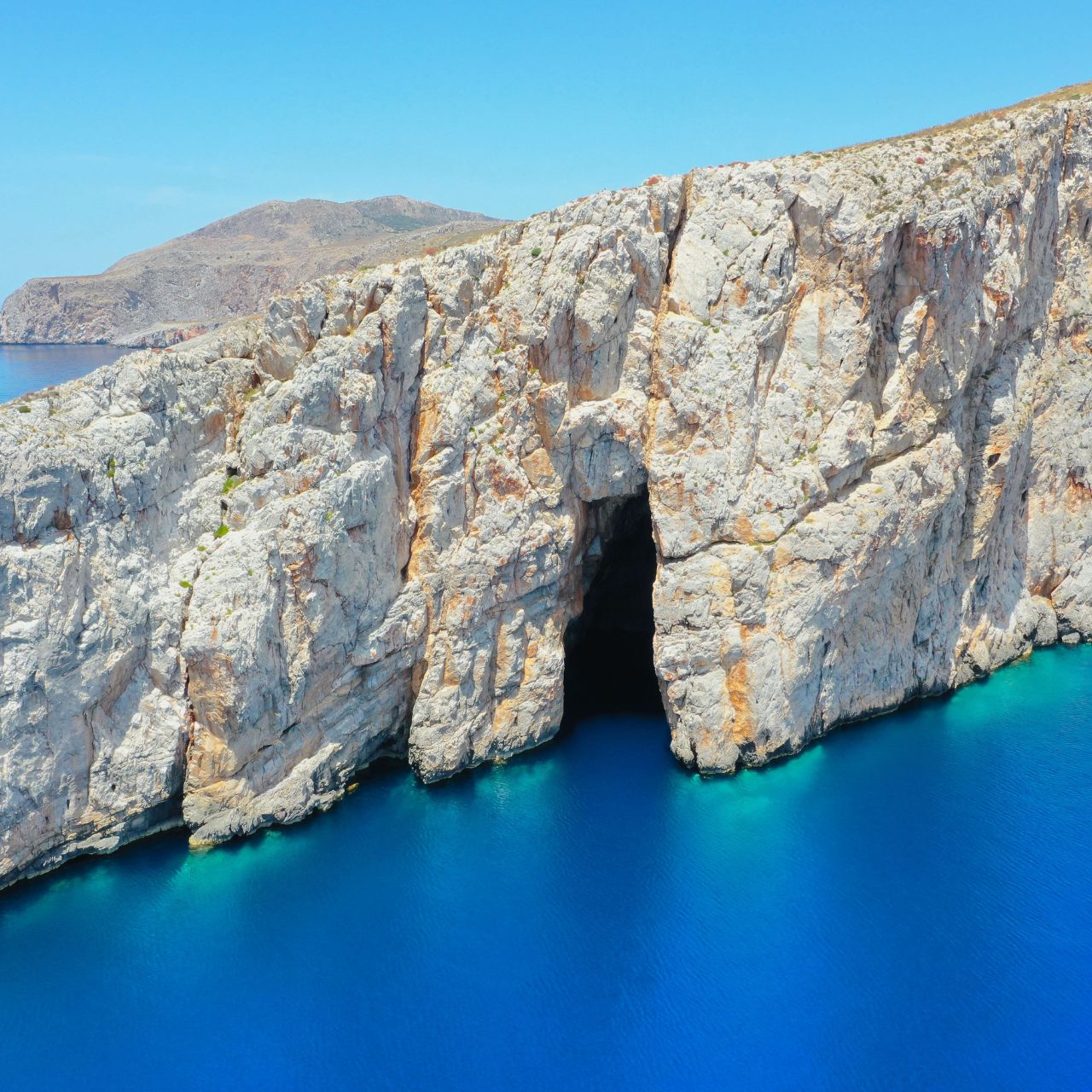 Cliffside rock formation along blue sea with a dark cave opening in the cliff edge under a clear sky