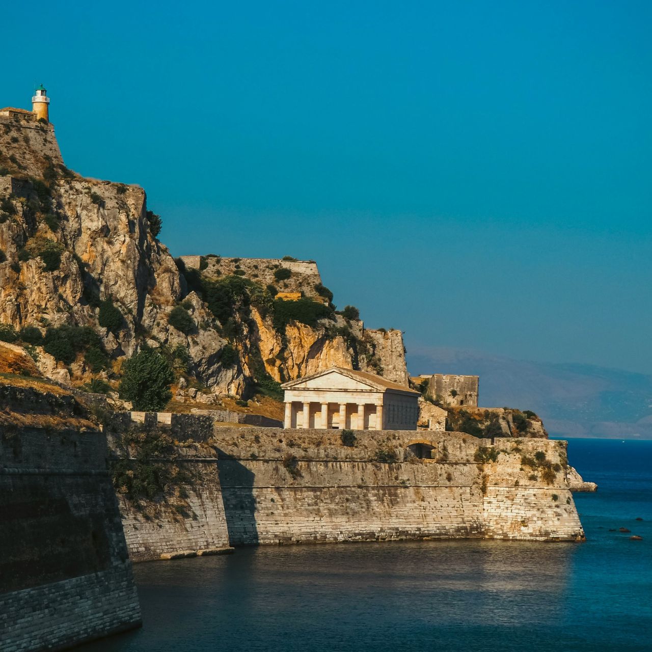 Cliffside fortress with a small lighthouse atop the hill and a classical temple-like building overlooking the sea.