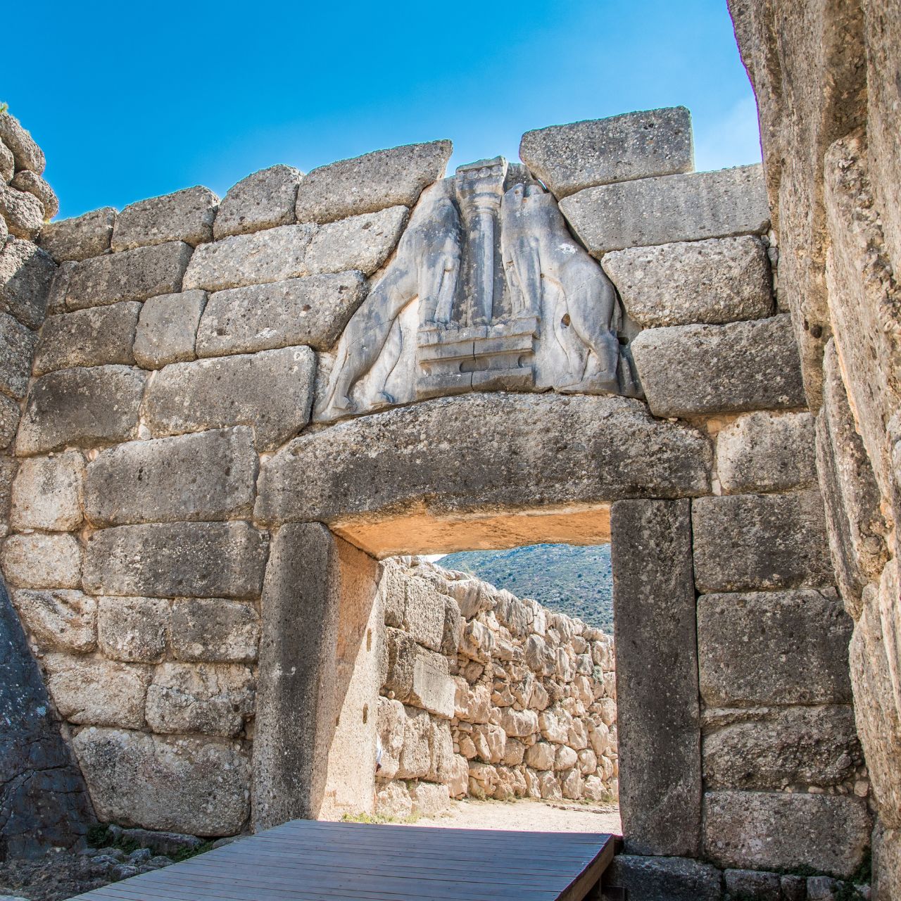 Ancient stone doorway in a dry-stone wall with a carved stone relief above the lintel, blue sky and distant hills beyond.