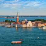 Aerial view of San Giorgio Maggiore with its tall bell tower and church, red-tiled buildings, and boats docked along the lagoon.