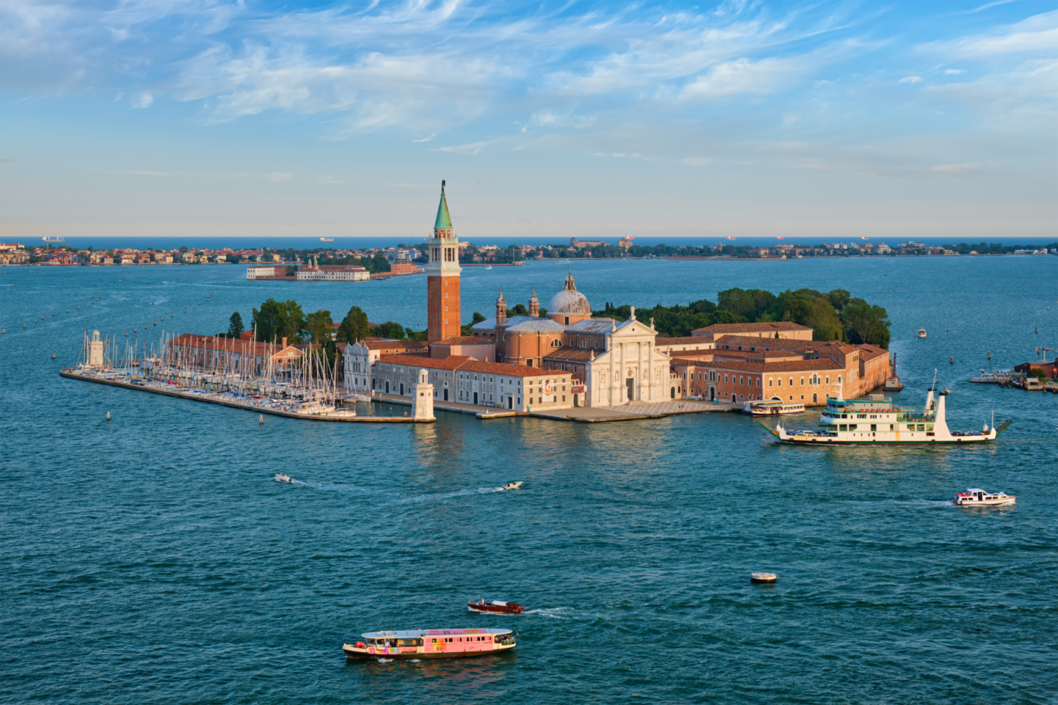 Aerial view of San Giorgio Maggiore with its tall bell tower and church, red-tiled buildings, and boats docked along the lagoon.