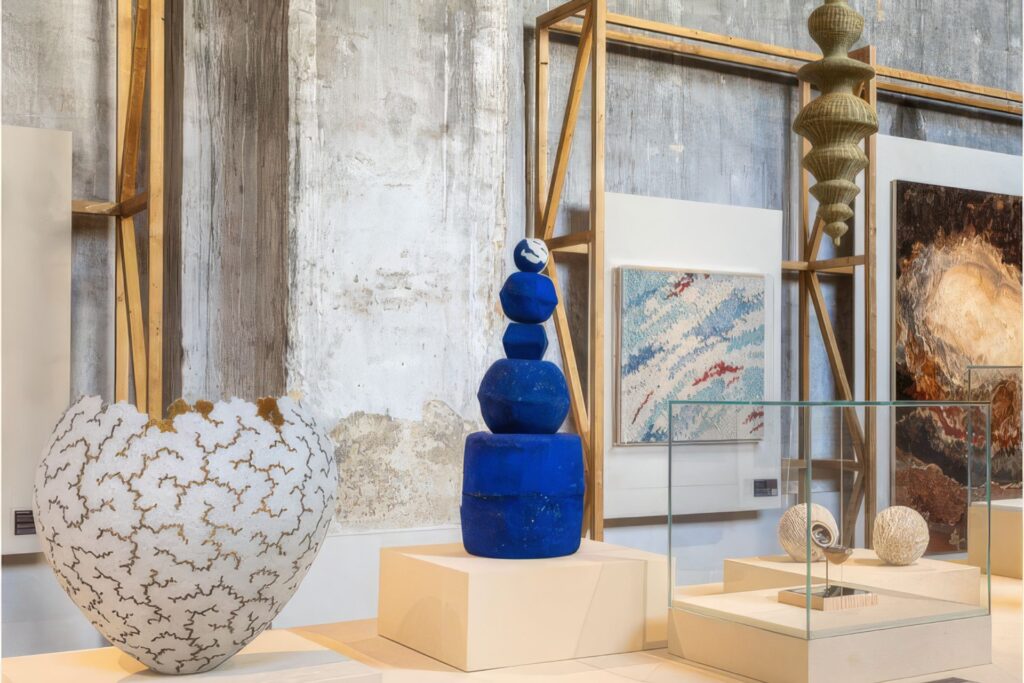 Art gallery interior with a blue stacked-stone sculpture on a pedestal, large white textured vase in the foreground, and framed artworks against a concrete wall.