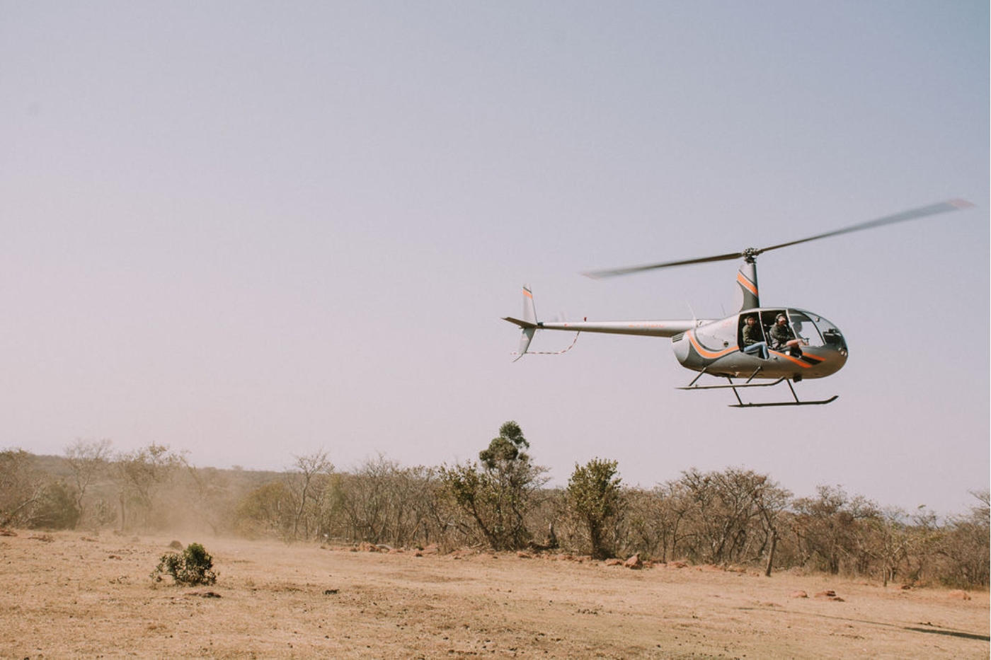 Light-colored helicopter hovering above a dusty desert landscape with two occupants in the cockpit.