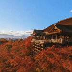 Traditional wooden temple perched on a platform above a sea of red autumn leaves with a city and hills in the distance under a blue sky.