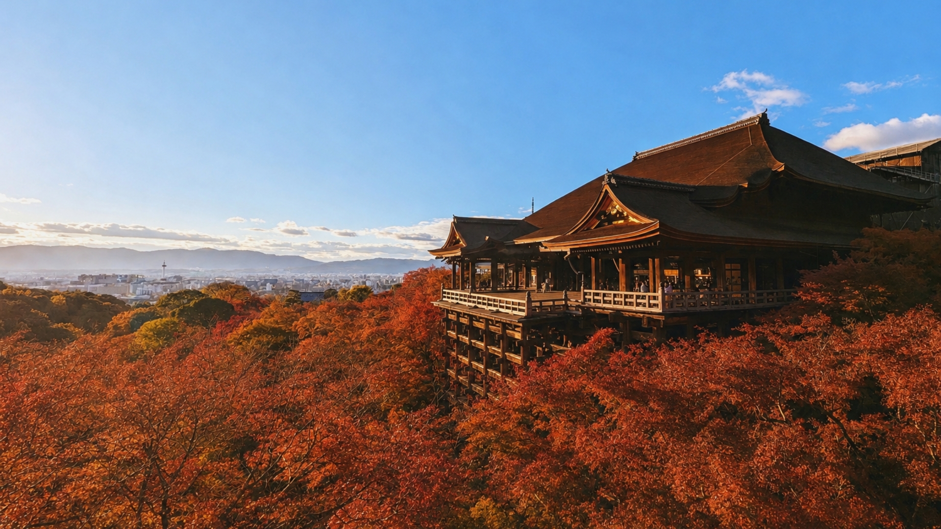 Traditional wooden temple perched on a platform above a sea of red autumn leaves with a city and hills in the distance under a blue sky.