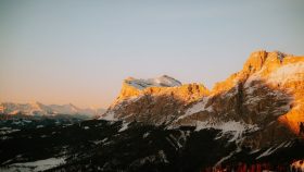 Snow-covered alpine peaks bathed in warm sunset light with a dark, forested valley below