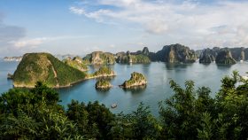 Calm blue bay with numerous green, rocky islands rising from the water under a bright sky; foreground frames with lush green foliage.