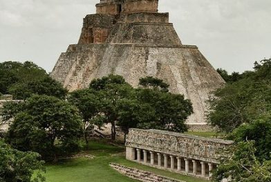 Ancient stepped pyramid rising above a grassy plaza, with a long colonnaded building in the foreground and trees around it.