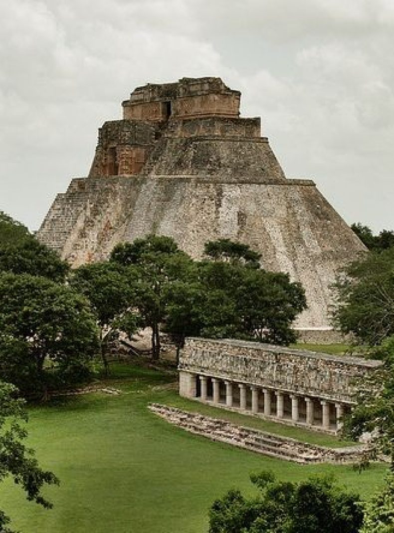 Ancient stepped pyramid rising above a grassy plaza, with a long colonnaded building in the foreground and trees around it.
